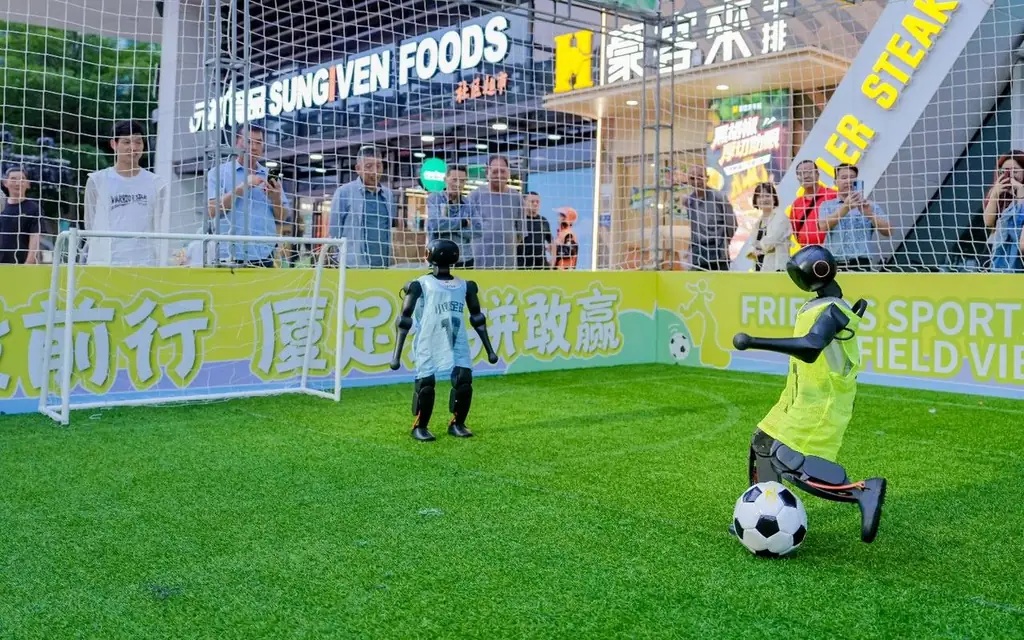 Two humanoid robots playing football inside a caged pitch at a shopping mall, with shoppers watching